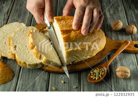 Hands expertly slice a golden-brown loaf of bread on a wooden board. Whole walnuts and spices rest nearby, adding a warm touch to this homely kitchen moment Hands expertly slice a golden-brown loaf of bread on a wooden board. Whole walnuts and spices rest nearby, adding a warm touch to this homely kitchen moment 122832935