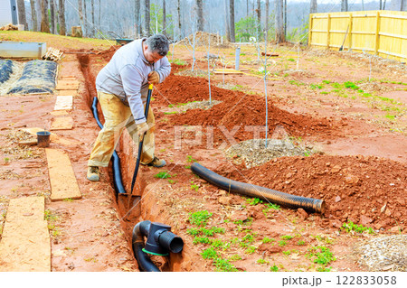 Contractor is digging trench to install drainage pipes in yard with newly planted trees on spring day. 122833058
