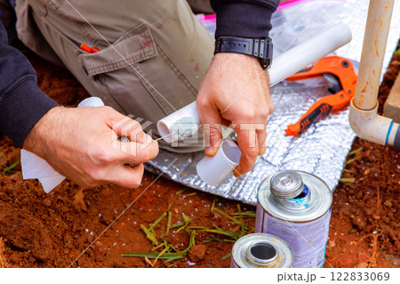 Worker applies adhesive to PVC pipe while kneeling on ground, surrounded by tools materials during repairs. Worker applies adhesive to PVC pipe while kneeling on ground, surrounded by tools materials during repairs. 122833069