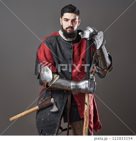 Medieval knight on grey background. Portrait of brutal dirty face warrior with chain mail armour red and black clothes and battle axe 122833154
