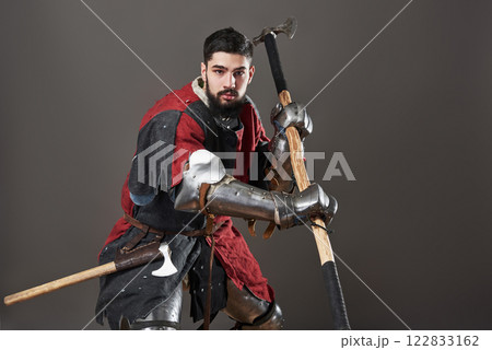 Medieval knight on grey background. Portrait of brutal dirty face warrior with chain mail armour red and black clothes and battle axe Medieval knight on grey background. Portrait of brutal dirty face warrior with chain mail armour red and black clothes and battle axe 122833162