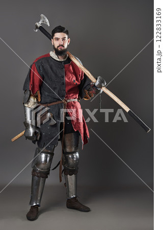 Medieval knight on grey background. Portrait of brutal dirty face warrior with chain mail armour red and black clothes and battle axe Medieval knight on grey background. Portrait of brutal dirty face warrior with chain mail armour red and black clothes and battle axe 122833169