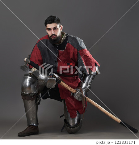 Medieval knight on grey background. Portrait of brutal dirty face warrior with chain mail armour red and black clothes and battle axe Medieval knight on grey background. Portrait of brutal dirty face warrior with chain mail armour red and black clothes and battle axe 122833171