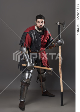 Medieval knight on grey background. Portrait of brutal dirty face warrior with chain mail armour red and black clothes and battle axe Medieval knight on grey background. Portrait of brutal dirty face warrior with chain mail armour red and black clothes and battle axe 122833175
