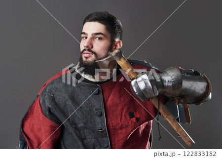 Medieval knight on grey background. Portrait of brutal dirty face warrior with chain mail armour red and black clothes and battle axe 122833182