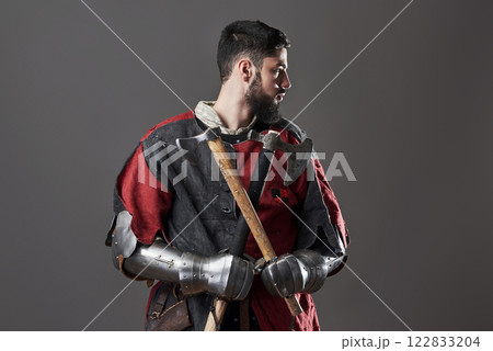 Medieval knight on grey background. Portrait of brutal dirty face warrior with chain mail armour red and black clothes and battle axe 122833204