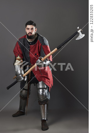 Medieval knight on grey background. Portrait of brutal dirty face warrior with chain mail armour red and black clothes and battle axe Medieval knight on grey background. Portrait of brutal dirty face warrior with chain mail armour red and black clothes and battle axe 122833205