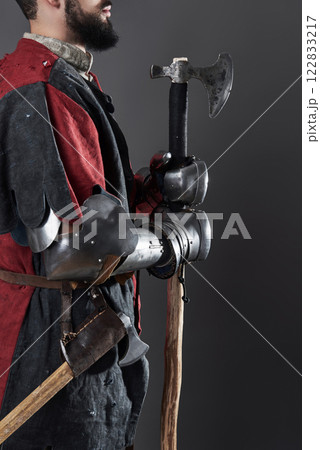 Medieval knight on grey background. Portrait of brutal dirty face warrior with chain mail armour red and black clothes and battle axe 122833217