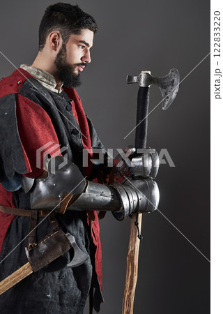 Medieval knight on grey background. Portrait of brutal dirty face warrior with chain mail armour red and black clothes and battle axe 122833220