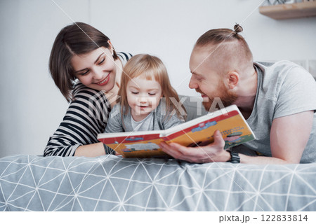 Father, Mother and Little Daughter Reading Children's Book on a Sofa in the Living Room. Happy big family read an interesting book on a festive day. Parents love their children Father, Mother and Little Daughter Reading Children's Book on a Sofa in the Living Room. Happy big family read an interesting book on a festive day. Parents love their children 122833814
