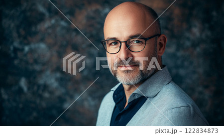 Man with glasses is smiling and wearing a blue and white sweater. He is standing in front of a wall 122834873