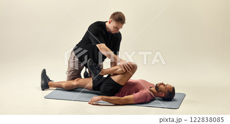 Trainer pressing bent knee while supporting lower back, helping with post-training recovery against beige studio background. 122838085