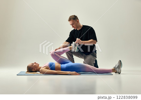 Male physiotherapist assisting woman lying on mat with leg stretch, supporting foot and knee to improve flexibility against light studio background. 122838099