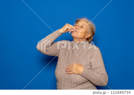 Caucasian woman in her 70s wearing a dark beige sweater licking her fingers after tasting a delicious delicacy. Isolated on blue background. 122840734