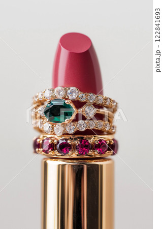 Pink lipstick surrounded by gold and gemstone rings on light background. Highlights beauty, luxury, and sophistication, perfect for glamour, makeup, cosmetics, fashion, jewelry-related themes. 122841693