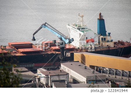 Industrial Port large Cargo Ship docked Freight Terminal. Blue Gantry Crane transferring goods from Vessel. Warehouses and calm Harbor water surface background, indicating busy maritime Logistics Hub Industrial Port large Cargo Ship docked Freight Terminal. Blue Gantry Crane transferring goods from Vessel. Warehouses and calm Harbor water surface background, indicating busy maritime Logistics Hub 122841944