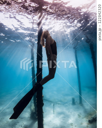 Freediver in swimsuit floats under the sunlit ocean surface, surrounded by light rays and underwater pillars, serene and free Freediver in swimsuit floats under the sunlit ocean surface, surrounded by light rays and underwater pillars, serene and free 122842320