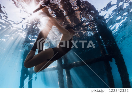 Freediver woman swims underwater under the pier in blue ocean with sun rays. 122842322