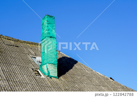 A house chimney covered with a green safety net on a gray damaged slate roof against a blue sky A house chimney covered with a green safety net on a gray damaged slate roof against a blue sky 122842788