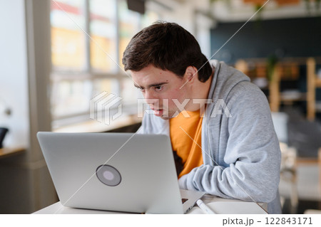 Concentrated young man with Down syndrome sitting and studying indoors at school, using laptop 122843171