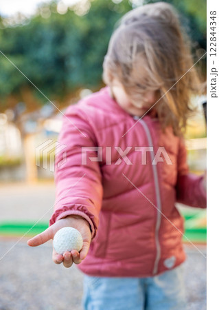 Portrait of Little girl Holding in hand Mini-Golf Ball. Little girl playing mini golf outdoor. Focus on the ball. Family vacations or resort. 122844348