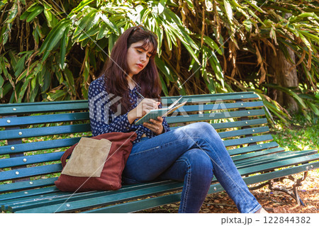 Young woman writing in notebook sitting on bench in park 122844382