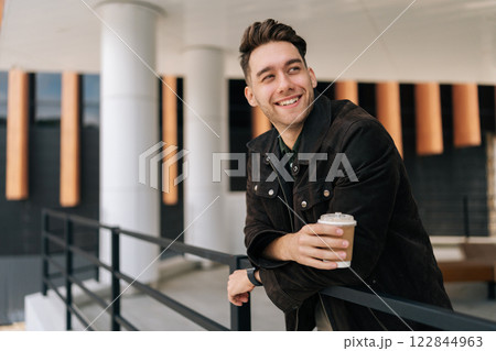 Handsome positive young man relaxing during coffee break, leaning on railing standing by modern office building. Happy bearded male in casual attire smiling looking away. Concept of urban lifestyle. Handsome positive young man relaxing during coffee break, leaning on railing standing by modern office building. Happy bearded male in casual attire smiling looking away. Concept of urban lifestyle. 122844963