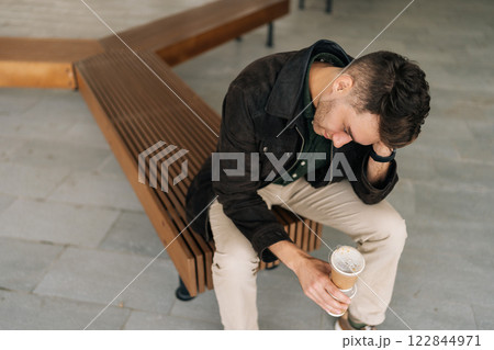 Pensive young man sitting on street bench, holding coffee cup and resting head on hand, lost in thought amid urban hustle and bustle of city life. Contemplative bearded male sitting on city bench. Pensive young man sitting on street bench, holding coffee cup and resting head on hand, lost in thought amid urban hustle and bustle of city life. Contemplative bearded male sitting on city bench. 122844971