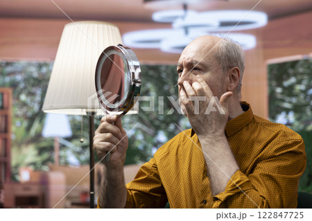 Elderly person using a mirror to apply grooming skincare products on his aging face, fighting against old age and wrinkles. Man doing his daily routine with UV protection cream and serum. Elderly person using a mirror to apply grooming skincare products on his aging face, fighting against old age and wrinkles. Man doing his daily routine with UV protection cream and serum. 122847725