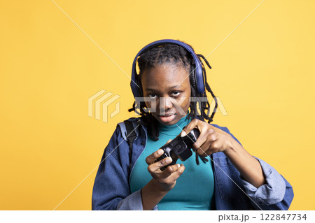 African american gamer holding controller, happily playing videogames, wearing headphones. BIPOC person excited to do gaming with gamepad, isolated over yellow studio backdrop African american gamer holding controller, happily playing videogames, wearing headphones. BIPOC person excited to do gaming with gamepad, isolated over yellow studio backdrop 122847734
