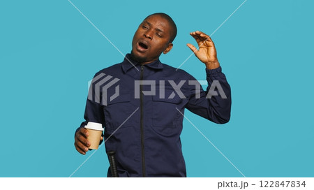 Portrait of fatigued security guard drinking coffee cup to remain energized while overseeing premises. Private law enforcement worker consuming caffeinated beverage, studio backdrop, camera A Portrait of fatigued security guard drinking coffee cup to remain energized while overseeing premises. Private law enforcement worker consuming caffeinated beverage, studio backdrop, camera A 122847834