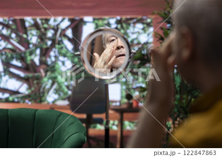Senior man looking at his aging face in the mirror and trying to preserve his skin, using hydration and UV protection serum. Elderly person applying grooming products on aging skin. 122847863