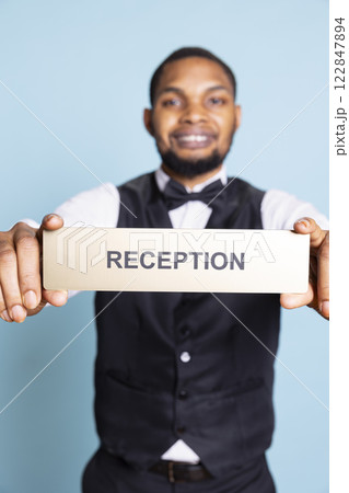 Skilled bellhop guiding clients to the reception using a wall sign at five star hotel, working in hospitality industry. Lodging personnel holding an indicator to assist guest during their stay. 122847894