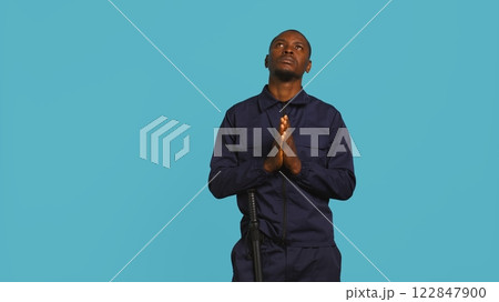 Portrait of security guard waiting for miracle, putting hands together while overseeing premises. Private law enforcement worker looking to sky, praying, studio backdrop, camera B 122847900