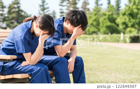 公園のベンチに座り落ち込むスクラブ姿の看護師・介護士・医療従事者の男女スタッフ 公園のベンチに座り落ち込むスクラブ姿の看護師・介護士・医療従事者の男女スタッフ 122849307