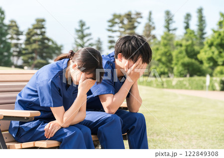 公園のベンチに座り落ち込むスクラブ姿の看護師・介護士・医療従事者の男女スタッフ 122849308