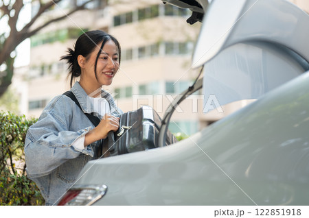 A smiling Asian woman places her luggage in the trunk of a car, getting ready for her trip. A smiling Asian woman places her luggage in the trunk of a car, getting ready for her trip. 122851918
