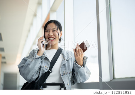 A cheerful Asian female passenger enjoys talking on the phone while waiting for her flight. A cheerful Asian female passenger enjoys talking on the phone while waiting for her flight. 122851952