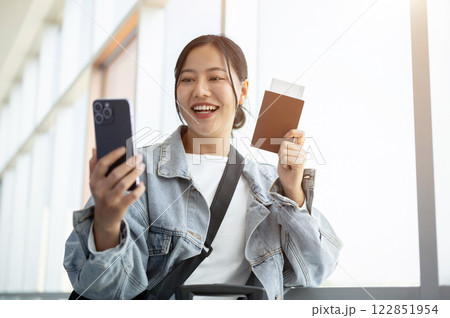 A cheerful Asian female passenger enjoys talking on a video call while waiting for her flight. A cheerful Asian female passenger enjoys talking on a video call while waiting for her flight. 122851954