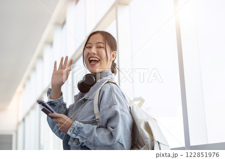 A cheerful Asian female traveler, waving to the camera while walking through the airport corridor. A cheerful Asian female traveler, waving to the camera while walking through the airport corridor. 122851976