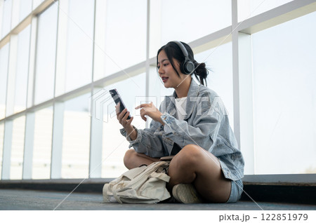 A beautiful Asian female passenger sits on the floor in the airport terminal, enjoying music. 122851979