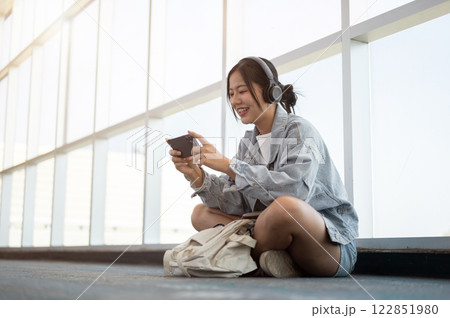 A cheerful Asian female passenger enjoys playing a mobile game while waiting for her flight. A cheerful Asian female passenger enjoys playing a mobile game while waiting for her flight. 122851980