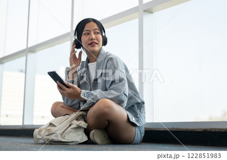 A beautiful Asian female passenger sits on the floor in the airport terminal, enjoying music. A beautiful Asian female passenger sits on the floor in the airport terminal, enjoying music. 122851983
