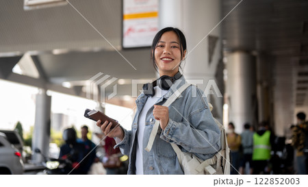 An attractive, smiling Asian female stands at an airport pickup point, waiting for a taxi. 122852003