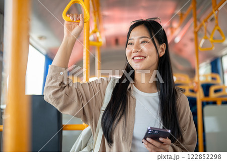 A cheerful Asian woman holds a handrail while admiring the city view during her bus ride. A cheerful Asian woman holds a handrail while admiring the city view during her bus ride. 122852298