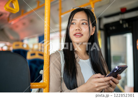 A beautiful Asian woman on a public bus leans against a pole, admiring the city landscape. 122852301