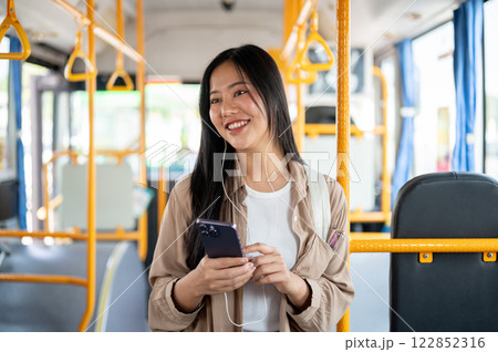 An attractive, cheerful Asian woman stands on a bus, admiring the city view during her ride. 122852316