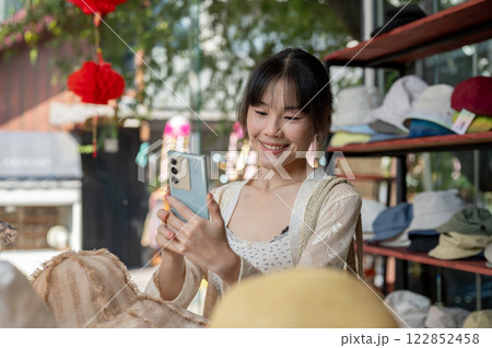 A happy, smiling Asian female customer is taking a picture of items in a hat shop, enjoying shopping A happy, smiling Asian female customer is taking a picture of items in a hat shop, enjoying shopping 122852458