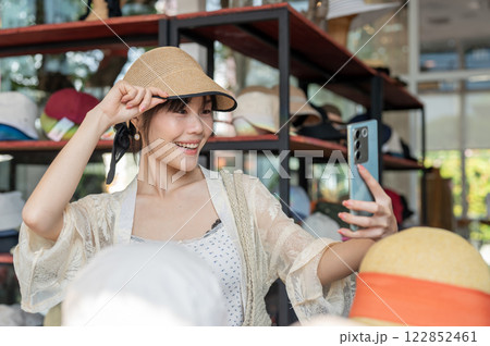 Cheerful Asian-Chinese female tourist enjoys blogging on her smartphone while shopping in a hat shop Cheerful Asian-Chinese female tourist enjoys blogging on her smartphone while shopping in a hat shop 122852461