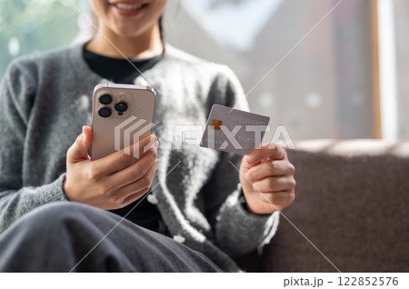 A close-up of a woman sits indoors on a bright day, holding a smartphone and a credit card. 122852576